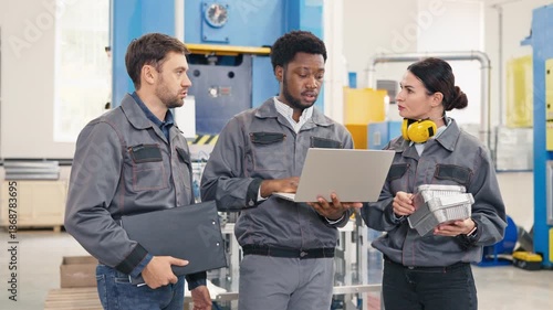 Male factory worker showing production data on laptop to colleagues. Woman holding foil containers checking quality. Man besides analyzing report. Team discussing manufacturing results in factory.
