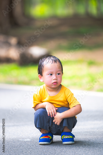 Candid vertical portrait of a curious young boy in a yellow shirt, crouching on a park path. The soft outdoor lighting and lush green bokeh background highlight a moment of innocent childhood discover
