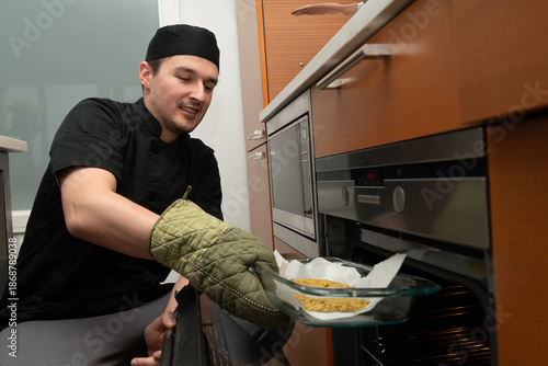 Professional chef places a dish into a modern oven while wearing an oven mitt and chef hat. The dish sits on parchment paper and is ready to be baked in this stainless steel appliance.