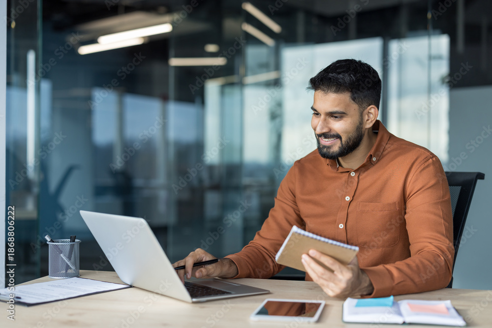 Obraz premium Young bearded indian male professional working on a laptop at a contemporary office desk, smiling while holding a notebook, and managing tasks with technology