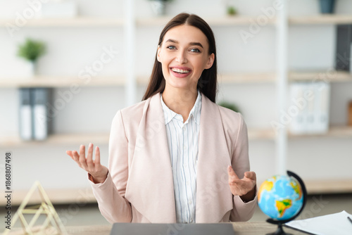 Foto Cheerful Female Travel Agent Talking To Camera Having Distant Consultation With Tourists Sitting At Desk In Tour Agency Office