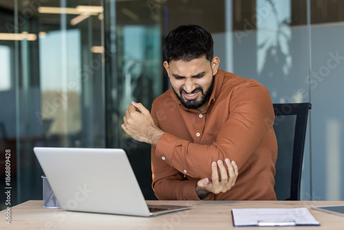 Photos Young indian businessman clutching his elbow in pain while working on a laptop a