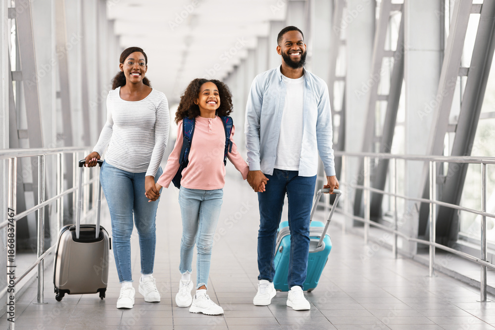 Fototapeta premium Family Travel Concept. Portrait of excited black dad, mum and girl walking at airport terminal corridor with suitcases waiting for the aircraft arrival, three people holding hands, free copy space