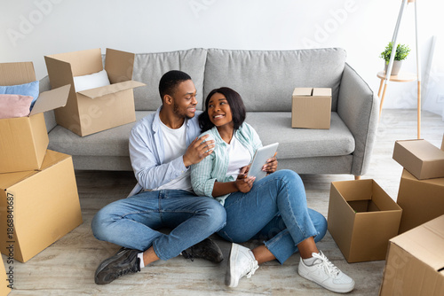 Valokuva Excited black spouses with touch pad searching household goods for their new home, using tablet on moving day, sitting on floor among boxes