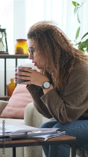 Vertical video of a young woman in glasses working on her laptop and drinking coffee while sitting on a sofa in her cozy living room.