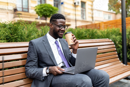 Tableau sur toile Corporate african american executive in formal wear sitting on bench near office center with laptop and takeout coffee during lunch break