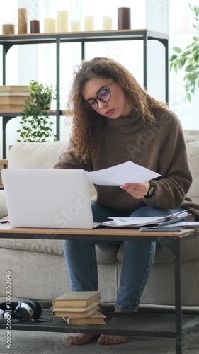A focused woman works from home on her laptop, sorting through documents and paperwork while sitting on the sofa in her living room. Vertical video.