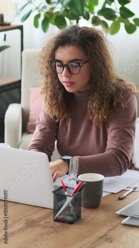 Vertical video of a thoughtful young woman working on a laptop, analyzing documents, and writing notes at her home office desk.