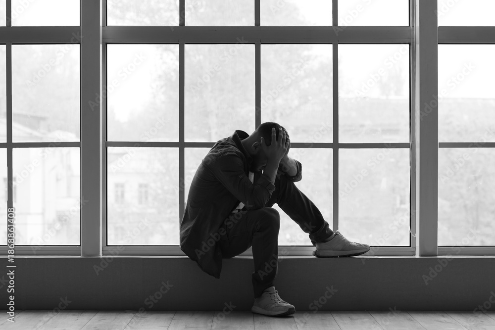 Fototapeta premium Depressed young man sitting near window at home. International Stress Awareness Week