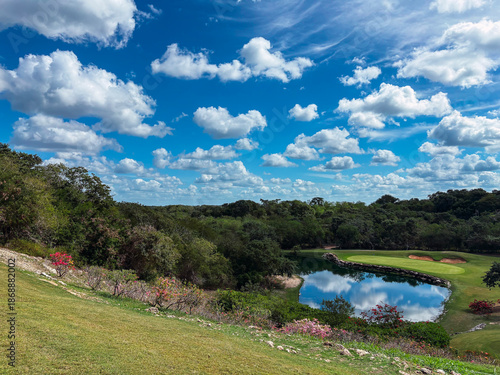 Beautiful landscape of a golf course, trees, grass, the pond, and some hills can be distinguished.
