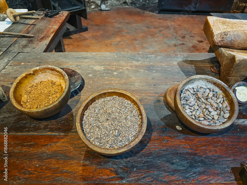 Ground cocoa beans on a rustic wooden table, accompanied by several clay pots with spices to prepare cocoa drink.

