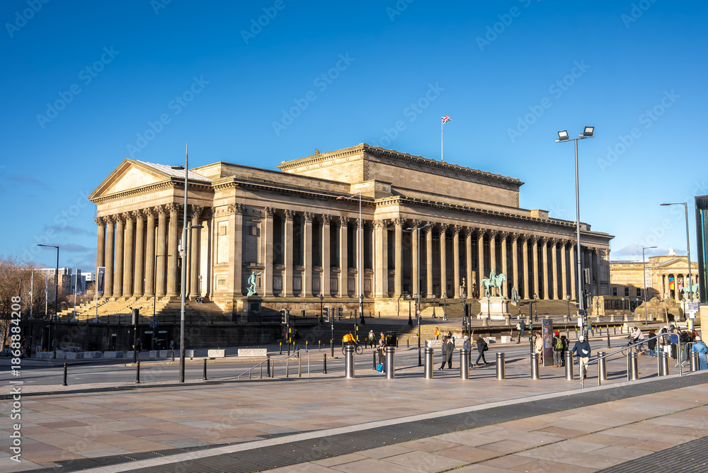 Fototapeta premium St George's Hall in Liverpool, England, shows a long colonnade, equestrian statues, and a Union Jack. People in winter coats cross the plaza under a crisp blue sky.