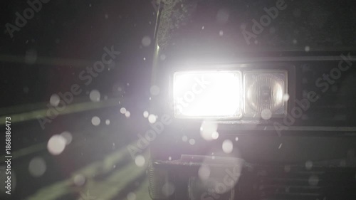 A close-up of a vintage vehicle s rectangular headlight glowing intensely in the dark. Snowflakes fall in the foreground, creating soft, blurry white bokeh circles against the moody night atmosphere.