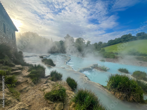 Natural turquoise thermal water terraces with rising steam and sun rays in Tuscany countryside.