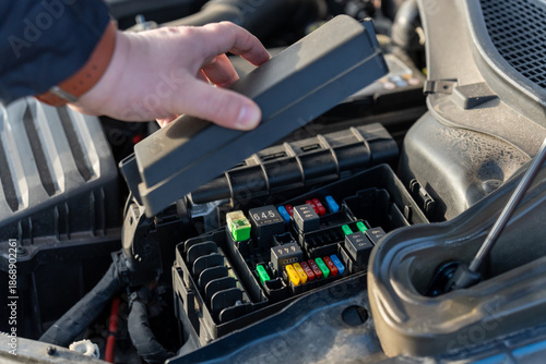 Male hand lifting plastic cover of automotive fuse box revealing colorful blade fuses and relays in car engine compartment. Concept of electrical system inspection, vehicle diagnostics and DIY repair.