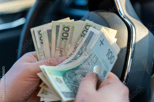 Close-up of hands counting fan of Polish zloty 100 banknotes near steering wheel inside car interior. Concept of cash payment, driver salary, vehicle expenses and personal finance management in Poland