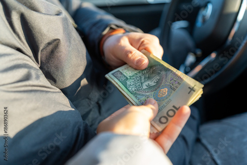 Passenger handing Polish zloty 100 banknotes to driver inside car as cash payment for taxi ride or vehicle transaction. Concept of transport service, fare payment, cash deal, personal money transfer.