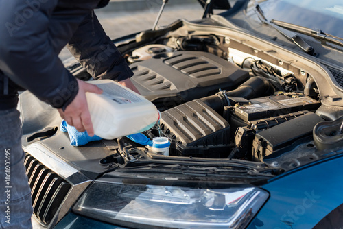 Man in dark jacket pouring windshield washer fluid from white canister into reservoir under open hood of modern car with visible grille. Concept of DIY car maintenance and winter preparation. 