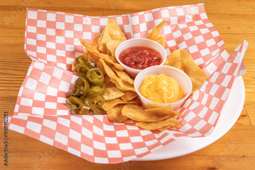 Mexican tortilla chips or nachos on red checked paper with jalapeno slices, salsa and cheese dip sauces on the side