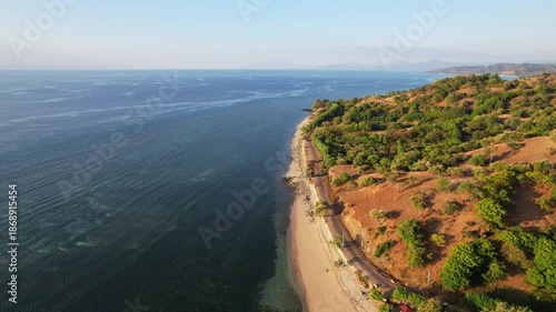Aerial drone footage of a white sand beach, with asphalt road in the front, and a flat panoramic blue seascape, dry bushes, orange sunset time, in Teluk Gurita, Belu Regency, Timor island, Indonesia