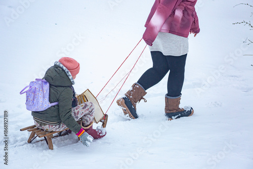 Family with parent holding a rope walking a child on a sled through field of snow seen from above. Winter wonderland ambient holidays fun street 