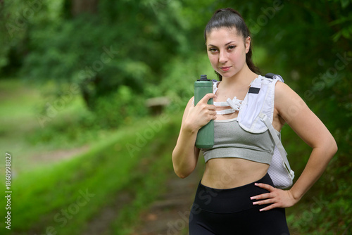 Beautiful young brunette female hiker pauses along a rail trail - holding green metal water bottle