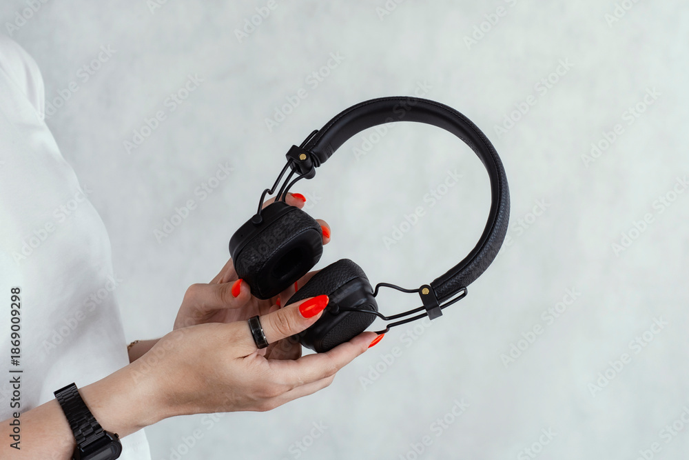 Naklejka premium Female hands with red manicure holding black wireless headphones against a light background. Music, audio technology, modern lifestyle and listening concept.