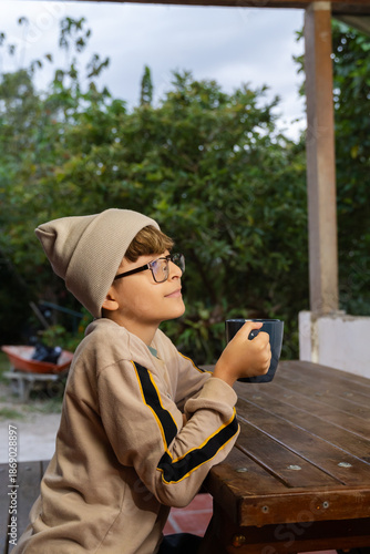 Young person holding mug at wooden table in backyard with foliage and wheelbarrow