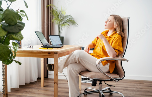 Slika na platnu Woman sitting in office chair with crossed legs looking at laptop and tablet