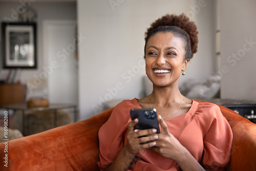 Fotografie Cheerful young Black smartphone user woman typing on cellphone, relaxing on soft