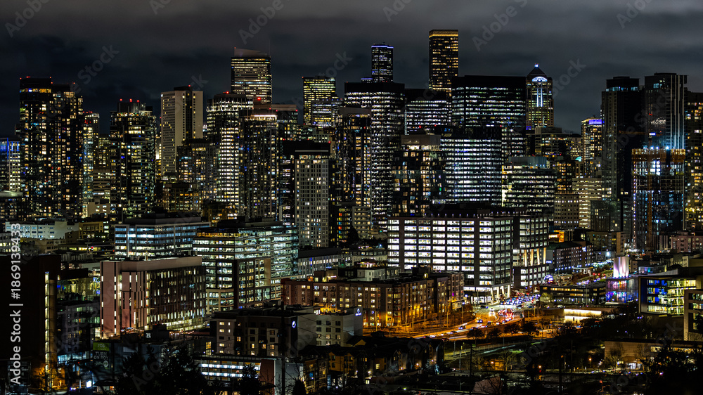 Obraz premium Night view of downtown Seattle showing dense illuminated skyscrapers, busy streets, and glowing windows under dramatic dark clouds. Ideal for business, technology, and urban lifestyle themes.