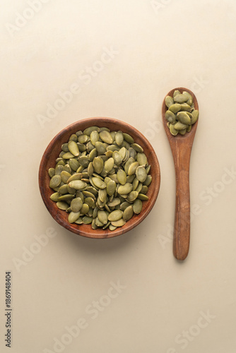 Green Pumpkin seeds in a wooden bowl placed on a beige background.