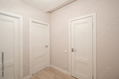 Neat hallway with three white paneled doors, light beige textured walls, light wood laminate floor, white ceiling, minimal decor, silver door handles, electrical outlets visible