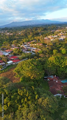 Aerial drone flight moving forward over the suburban residential area of San Rafael de Heredia, Costa Rica, with lush green mountains and a vibrant sunset