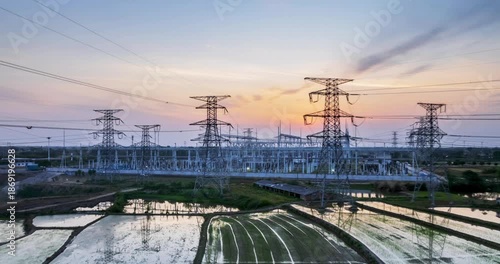 Aerial time-lapse of a large substation at dusk, power infrastructure amid golden-hour glow and reflective rice fields.