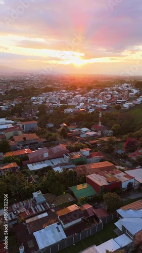 Vertical drone flight moving forward over San Rafael de Heredia, Costa Rica, with a view of the local church and San Jose city skyline during a golden tropical sunset