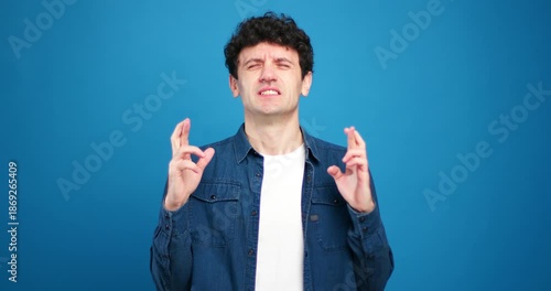 Handsome man with crossed fingers looking at camera having wish and dream on blue background