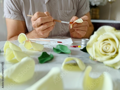 Bright conceptual photo of a muscular bearded man painting, gluing and assembling a flower made of air dry clay. Creative handmade process and art workshop concept, horizontal, focus on flower