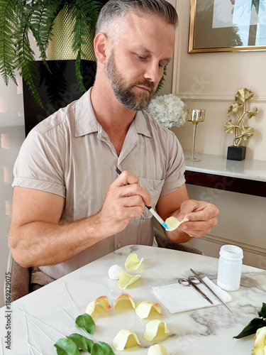 Bright conceptual photo of a muscular bearded man painting, gluing and assembling a flower made of air dry clay. Creative handmade process and art workshop concept, Vertical, focus on men
