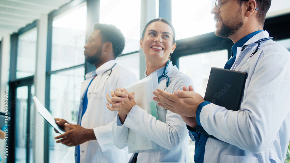 Fototapeta premium Cute female doctor smiling and clapping in bright hospital corridor. Male colleagues joining applause. Team celebrating success and appreciating strong performance during busy shift.
