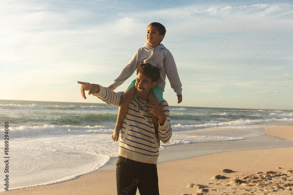 Fototapeta premium Happy father pointing to son while carrying him on shoulders at beach against sky