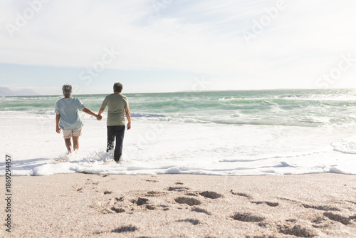 Rear view of senior multiracial couple holding hands wading on shore at beach during sunny day