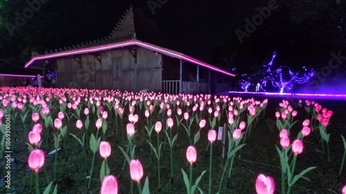 Nighttime scene of a warmly lit house with a decorative garden featuring soft pink artificial tulip lights and sparkling trees adorned with ornamental lighting, creating a magical atmosphere.