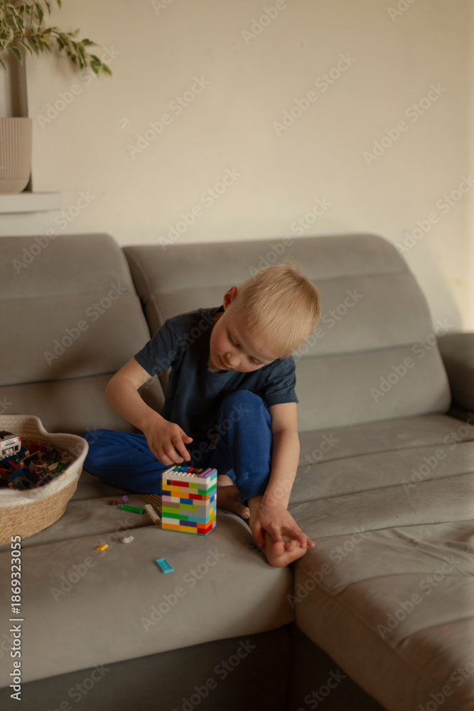 Naklejka premium White boy building colorful block tower on sofa, focused hands arranging bricks, woven basket of toys nearby, warm daylight, calm