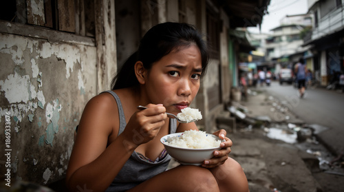 Young Filipina woman sits on a narrow urban street, holding a bowl of white rice and a spoon with a serious, direct gaze toward the camera.