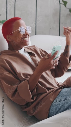 Red haired African American woman with glasses uses smartphone with surprised expression to message on screen.  Lifestyle sitting on sofa in home interior wearing casual sweater and jeans reacting 