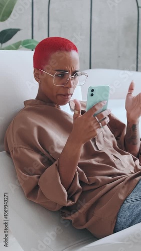 Red haired African American woman with stylish eyewear wearing clothes, using smartphone scrolling messages and apps, upset by message received, while relaxed pose on couch at home