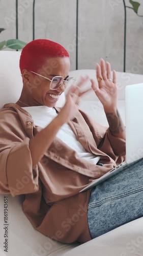 African American woman with red hair smiles while using a laptop and appears happy, sitting on a couch in a bright room. The background shows plants and a modern interior design