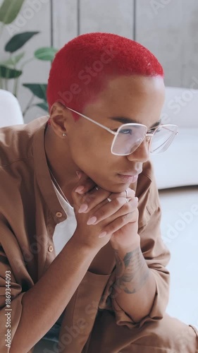 A person with red hair sits quietly in a bright room. They are lost in thought with their hands clasped and show a serious expression. The setting has plants and a simple design.