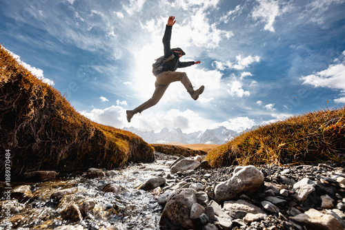 Man hiker with backpack jumps over small creek against high snow capped mountains. Active vacations concept